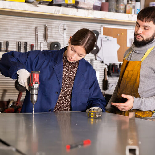 Eine junge Frau bohrt in einer Werkstatt während ein junger Mann anleitend daneben steht.