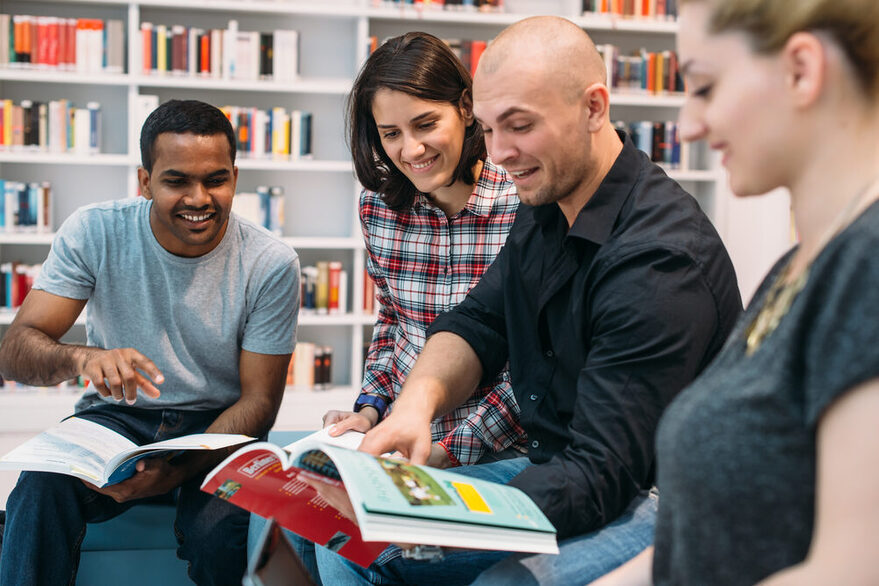 Vier Studenten sitzen in einer Bibliothek und betrachten fröhlich ein Buch.