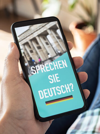 Ein Hand hält ein Smartphone. Auf Bildschirm steht "Sprechen Sie Deutsch?"