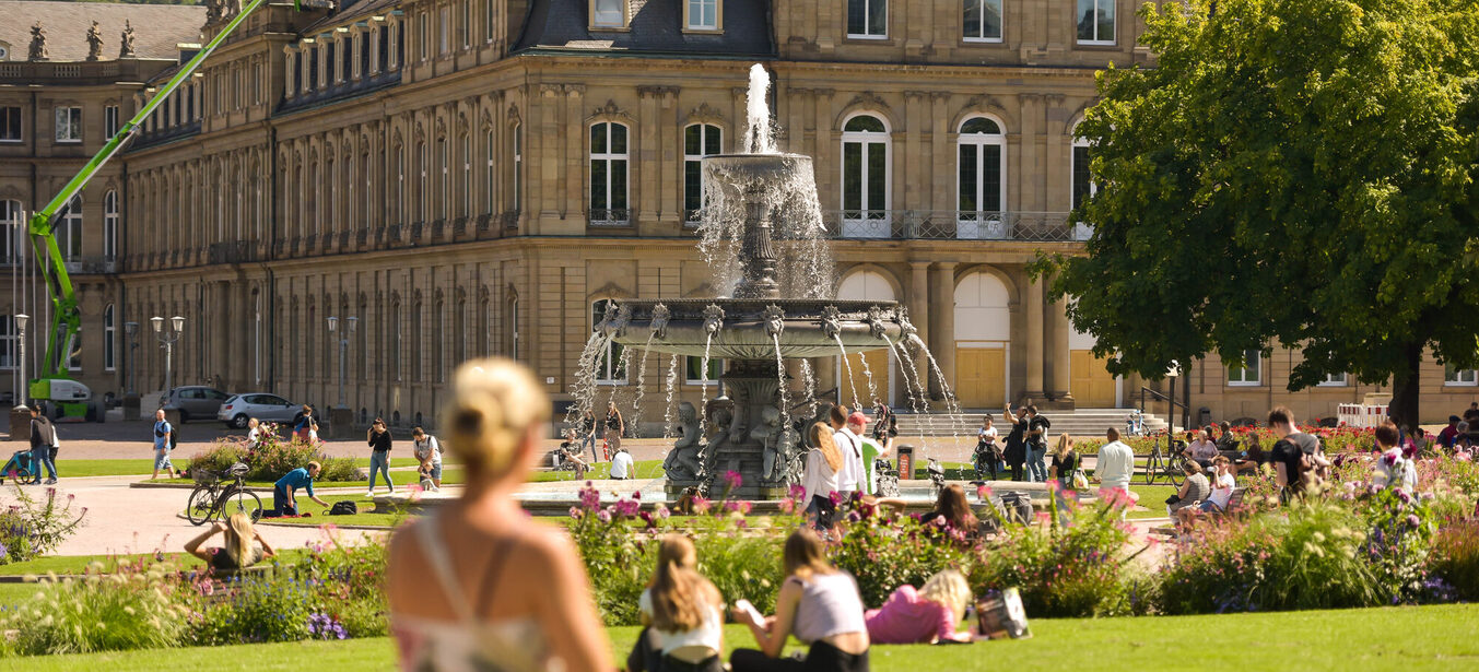 Am Schlossplatz sitzen Menschen bei Sonnenschein auf der Wiese und am Brunnen.