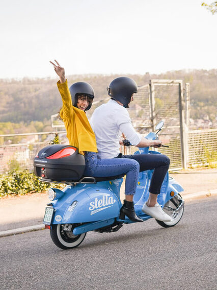 Ein Mann und eine Frau fahren mit einem blauen Roller auf einer Straße, die Frau grüßt in die Kamera.