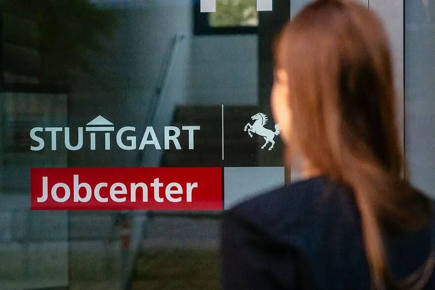 Der Eingang zum Jobcenter Stuttgart. Zu sehen ist der Schriftzug auf einer Glastür und eine unscharfe Person, die vor der Tür steht.